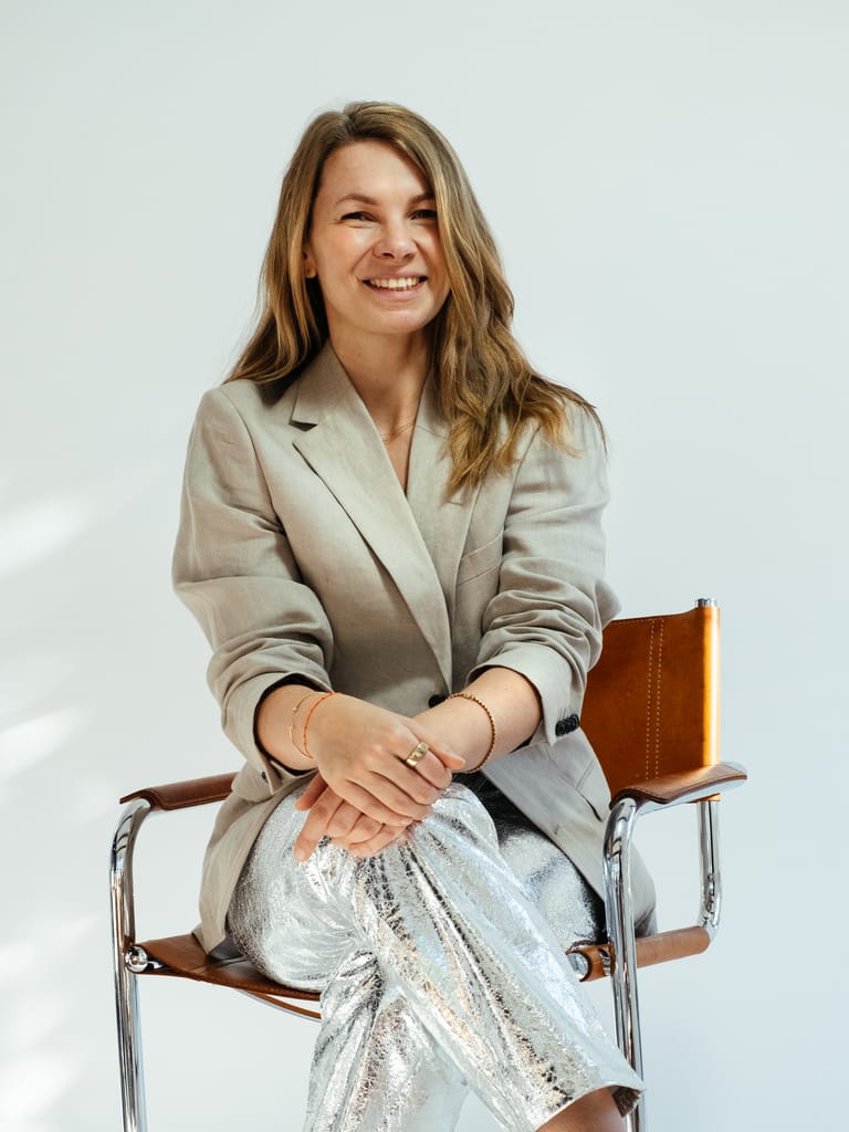 Smiling founder Carolin Moehring in beige blazer and silver metallic pants sits cross-legged on mid-century leather chair in bright studio.