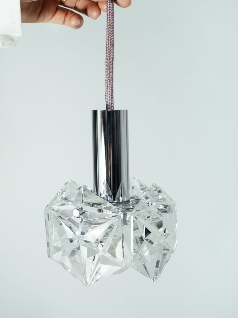 Hand holds mauve fabric cord suspending faceted crystal Kinkeldey Mémoire pendant with polished chrome stem against clean background.