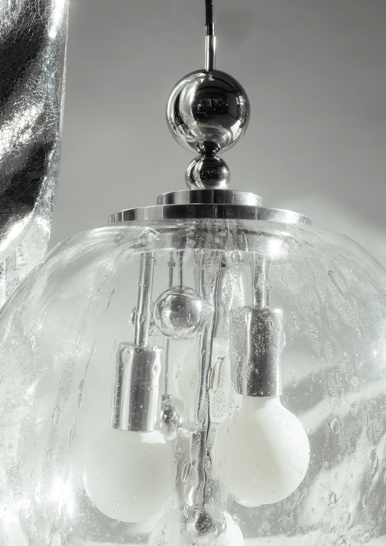 Close-up of mid-century chrome pendant Doria & Luna lamp inside textured glass globe, droplets framing white bulbs against soft grey.