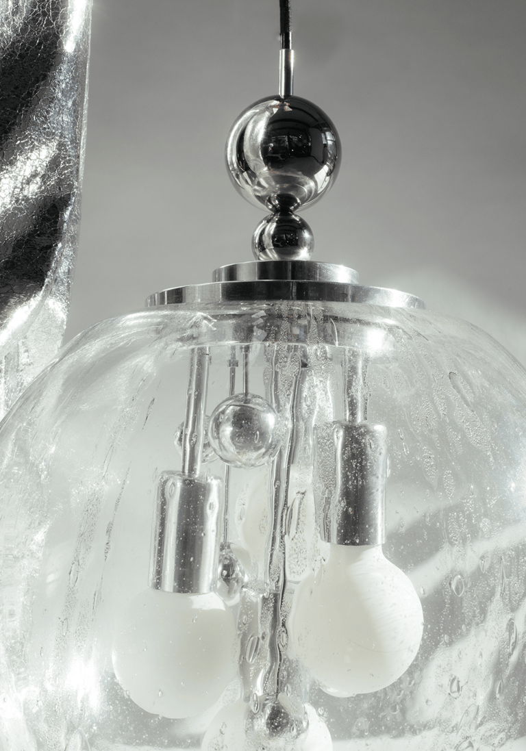 Close-up of mid-century chrome pendant Doria & Luna lamp inside textured glass globe, droplets framing white bulbs against soft grey.