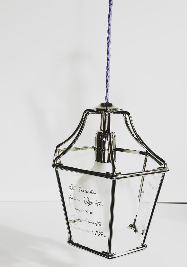 Harriet lantern pendant with handwritten German text on glass, hung by lilac twisted cord against stark white wall.