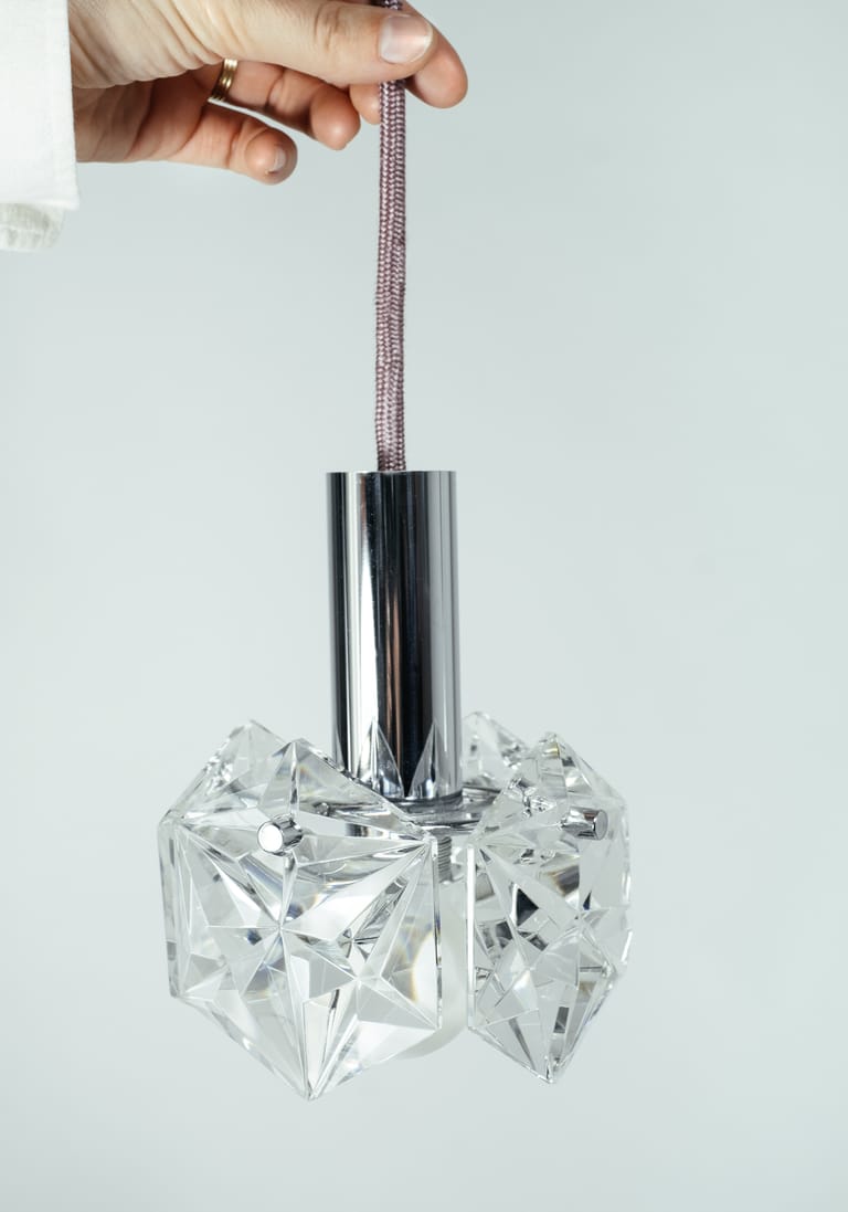Hand holds mauve fabric cord suspending faceted crystal Kinkeldey Mémoire pendant with polished chrome stem against clean background.