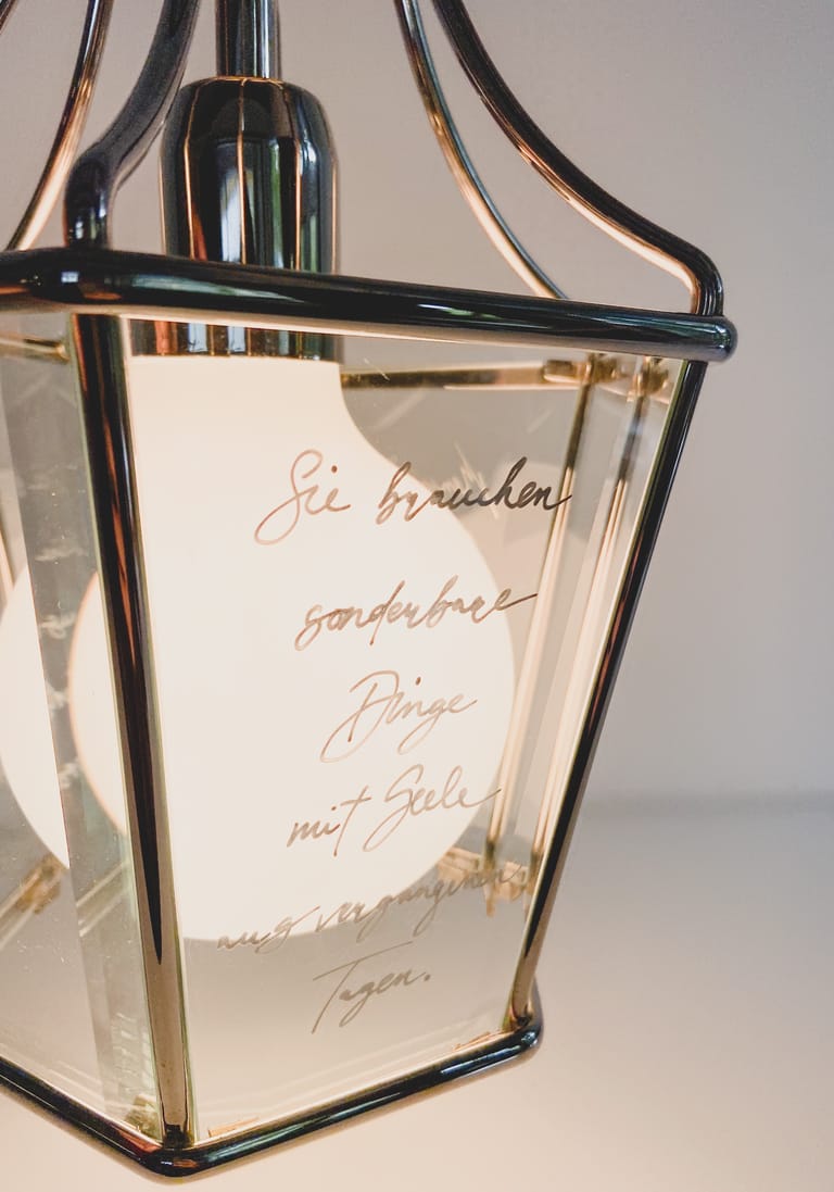 Detail view: handwritten German quote etched on glass of chrome Harriet lantern, warm globe glowing within.