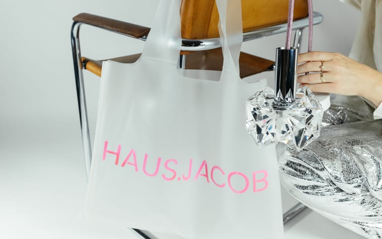 Hand holds crystal pendant beside frosted HAUS.JACOB bag in neon-pink lettering draped over chrome mid-century leather chair.