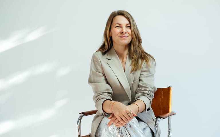 Smiling company founder and CEO in beige blazer and silver metallic trousers sits relaxed on leather chair in bright studio.
