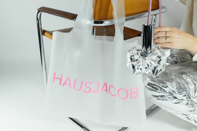 Hand holds crystal pendant beside frosted HAUS.JACOB bag in neon-pink lettering draped over chrome mid-century leather chair.