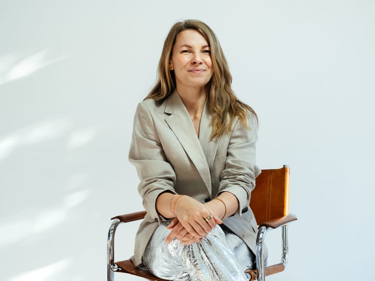 Smiling company founder and CEO in beige blazer and silver metallic trousers sits relaxed on leather chair in bright studio.