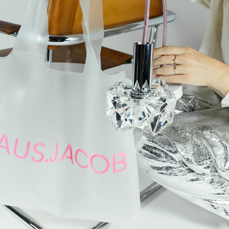 Hand holds crystal pendant beside frosted HAUS.JACOB bag in neon-pink lettering draped over chrome mid-century leather chair.