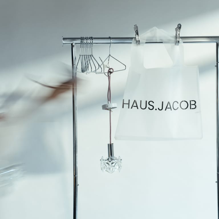 Blurred hand (from founder Carolin) sweeps chrome rack holding crystal pendant and frosted HAUS.JACOB bag, hangers gleaming in soft studio light.