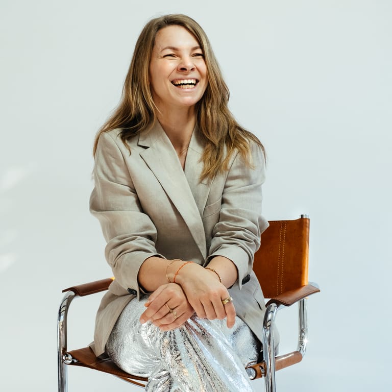 Laughing CEO Carolin Moehring in beige blazer and silver metallic trousers sits cross-legged on vintage leather chair in bright studio.